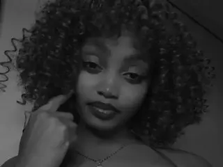 SinfulMuseTV poses close-up, showing her face and curly hair, wearing a thin necklace against a dark background.