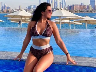 SireniaBaby poses in a brown patterned bikini, showing her cleavage and curves against a blue water background.