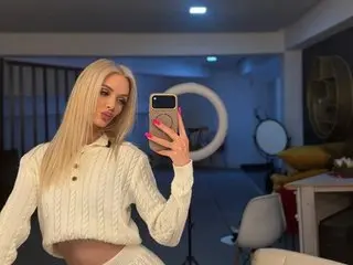 SkinnyNatty poses for a mirror selfie, wearing a white knit sweater and showing her long blonde hair and light skin.