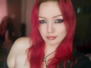 Portrait of a woman with vibrant red hair and blue eyes wearing dark makeup in a soft indoor setting.