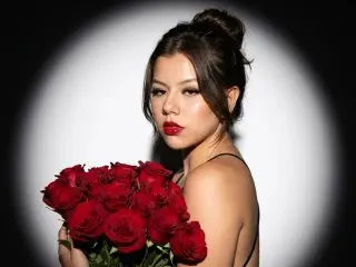 SkyValentiinee poses with a large bouquet of red roses against a spotlight background, looking at the camera with red lipstick.