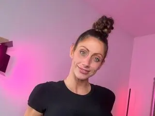 Smiling woman in a black t-shirt with hair in a bun against a pink lit background.