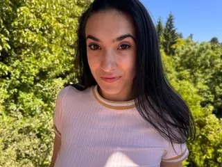 SlimTemptation poses with her long black hair down, looking directly at the camera against a bright, leafy green outdoor background.