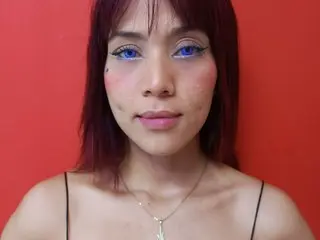 Young woman with red hair and blue eyes wearing a black camisole against a vibrant red background.