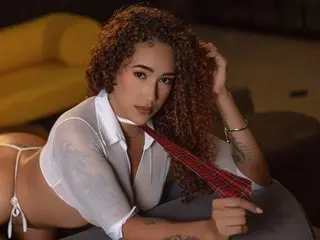 SofiaaV poses in a white button-down shirt and red tie, showing her curly hair and bare shoulders in a dim setting.