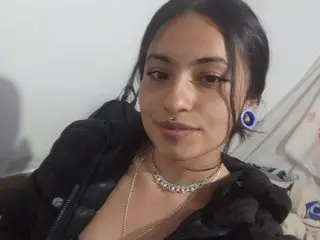 Sofisexxx69 poses close-up, wearing a black jacket and a silver necklace, showing her face and shoulders against a plain white wall.