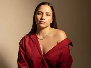 SonjaBela poses with long dark hair, wearing an open red button-down shirt that shows her cleavage against a plain tan background.