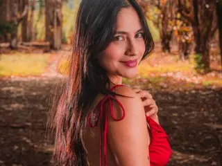 SophiaCastro32 smiles while looking over her shoulder, showing her long dark hair and red strap top against a soft, natural outdoor background.
