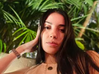 Young woman with long dark hair wearing a beige top in a sunny tropical garden setting.