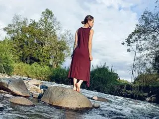 Sophiamiller18 stands outdoors in a long, sleeveless maroon dress, looking over her shoulder against a backdrop of green trees and a blue sky.