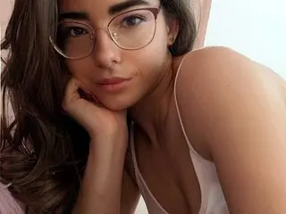 SophieVoss poses with her hand under her chin, showing her face, long brown hair, and white tank top strap against a plain background.