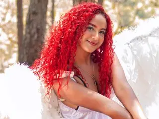 Sophie_Velvett poses with her long red curly hair, wearing a white top against a soft, sunlit forest background.