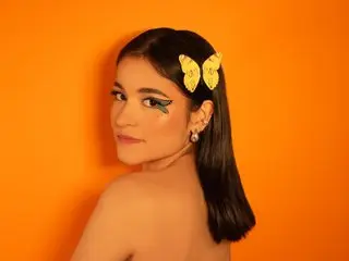SophieeJacobs poses with a side profile, showing her long dark hair and a yellow butterfly clip against a bright orange background.