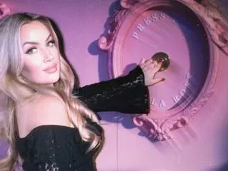 SouthernHoneyX poses with her long blonde hair and a black off-the-shoulder top against a purple background.