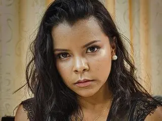 Serious young woman with long wavy dark hair wearing a black lace top in a warm indoor setting.