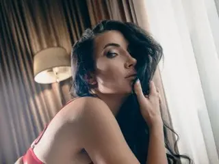 StassiLyne poses with her hand near her face, showing her dark wavy hair and bare shoulders against a dark, draped background.
