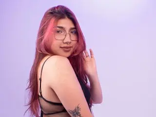 Young woman with pink hair and glasses wearing a black lace top against a soft purple background.