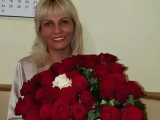 Smiling blonde woman in a white blouse holding a large bouquet of red roses in a bright indoor setting.