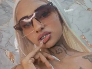 Sumaya69 wearing dark sunglasses and posing with her finger near her lips against a marble background.