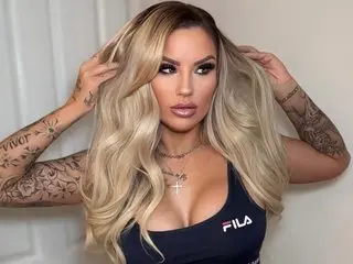 Summer_FoxxX with long blonde hair and tattoos posing in a black FILA tank top.