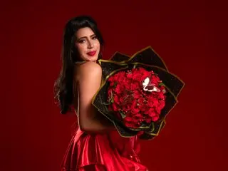 SummersAngel poses with long dark hair and red lipstick, wearing a red satin outfit while holding a large bouquet of red roses against a red background.