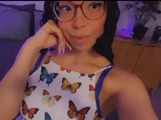 SupaNova777 wearing a white tank top with a colorful butterfly print, looking at the camera with her hand near her face.