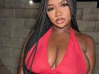 SupaNovaa poses in a red low-cut top, showing cleavage, with her long dark braids hanging down over her shoulders.