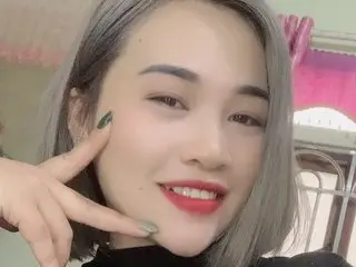 SuryBee poses with a hand near her face, showing her short blonde hair and red lipstick against a soft pink background.