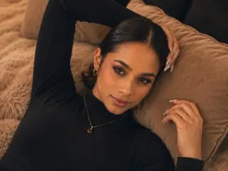 SusanBells poses with her hands behind her head, wearing a black long-sleeve turtleneck and showing her dark hair and face.