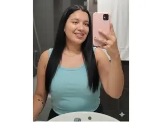 SusanStewart poses for a mirror selfie wearing a tight light blue tank top, showing her long black hair and smiling face.