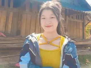 SusuBee smiles while wearing a yellow tank top and a blue and white jacket, with her hair pulled back in a messy bun.