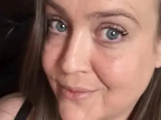 SuzyJohnston looks directly at the camera with a slight smile, showing her face and light brown hair against a dark background.
