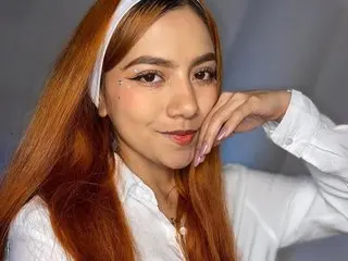 Young woman with long reddish hair and white headband wearing a white shirt in a soft, bright indoor setting.