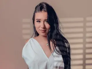 Young woman with long dark hair wearing a white blouse in a soft, sunlit indoor setting.