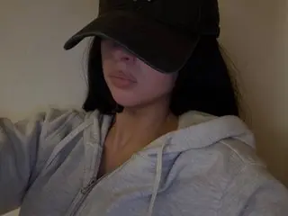 SweetScarletteUK wearing a black cap and a grey zip-up hoodie, looking forward with her long dark hair down.