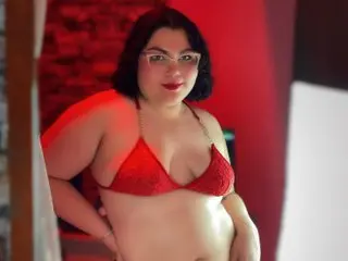 Sweet_Nova poses in a red lace bra, showing her cleavage and bare stomach against a red background.