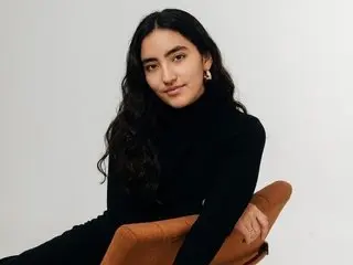 Sweetie_Angel with long dark hair wearing a black turtleneck sitting in a brown chair against a white background.