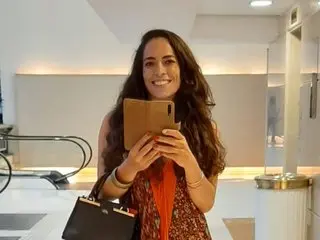 SweetsAnne smiles while holding a smartphone, showing her long wavy hair and a patterned orange top in a bright room.
