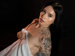 Tattooed woman in a white silk garment posing against a dark background with moody lighting.