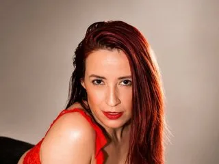 SweettEve — SweettEve poses with a direct gaze, her long red hair falling over her shoulders against a plain background.