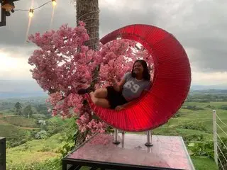 Sweettei sits smiling in a large red crescent moon chair surrounded by pink flowers overlooking a green valley.