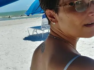 SwimStar poses on a sunny beach, showing her face, short dark hair, and bare shoulders against a blue beach umbrella and white sand.