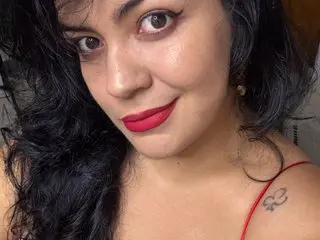 Smiling woman with dark wavy hair and red lipstick wearing a red strap outfit in a warm indoor setting.