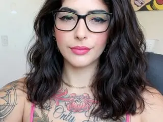 Woman with wavy dark hair, black glasses, and pink lipstick showcasing colorful chest tattoos in a bright setting.