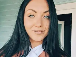 TanyaDevon poses with long, straight black hair and a direct gaze, showing her face and shoulders against a light-colored wall.