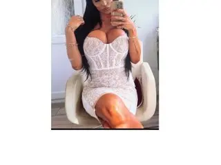 TaraJames99 poses in a white lace dress while sitting in a white chair and taking a mirror selfie.