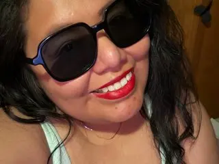 TaraWinter smiles while wearing dark sunglasses and a white tank top, showing her long dark hair and red lipstick.