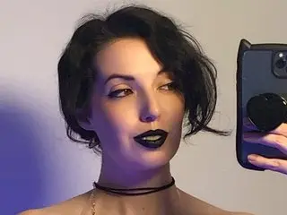 TashaNoir poses with a direct gaze, wearing a black choker and dark lipstick with her black hair styled in a short bob.