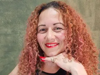 TatianaBlonde smiles with her long, curly reddish-blonde hair, wearing a black top against a plain green background.