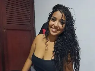 Tatihot69 smiles while posing with her long, curly dark hair, wearing a black tank top top in an indoor setting.